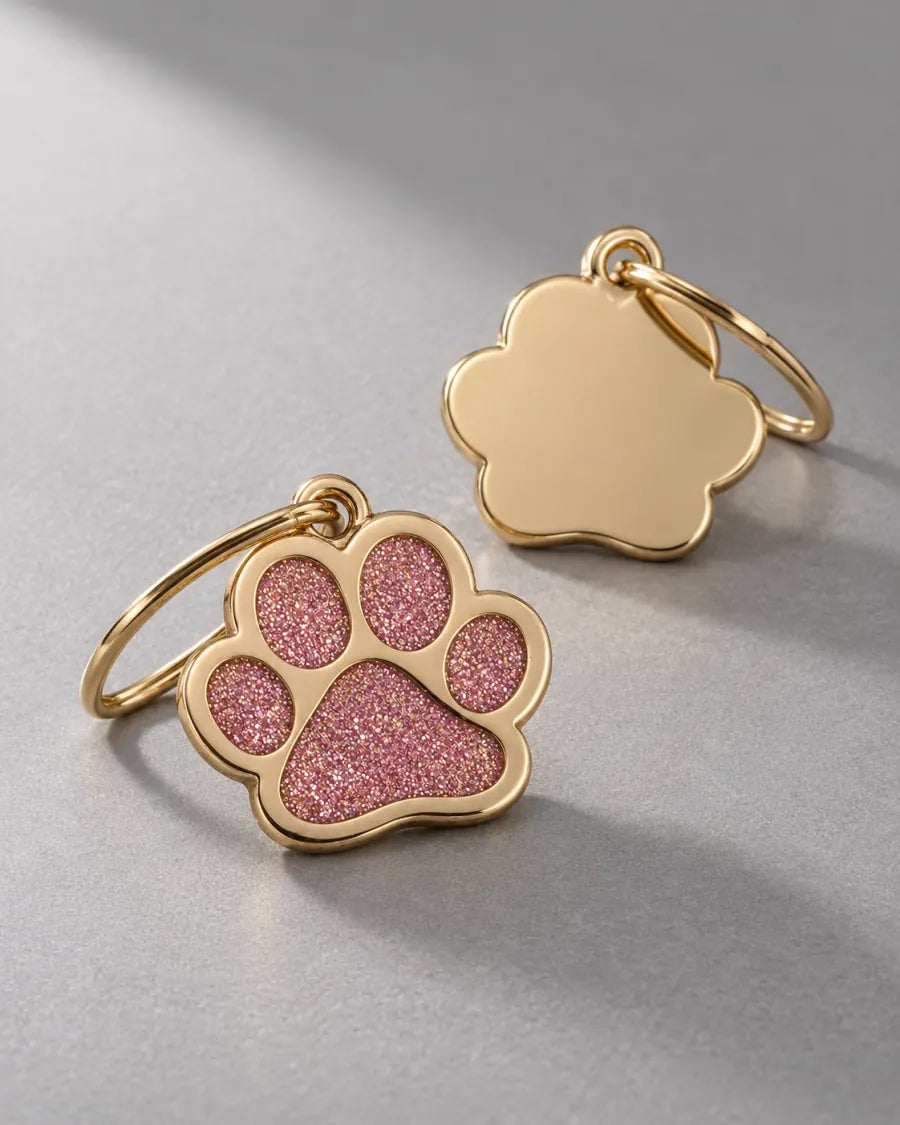 Gold paw-shaped pet tags with pink glitter pads photographed in a clean cool-grey studio